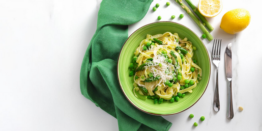 A green bowl of long pasta with peas and asparagus. Next to the bowl is a green tea towel, cutlery, lemon, peas and asparagus.