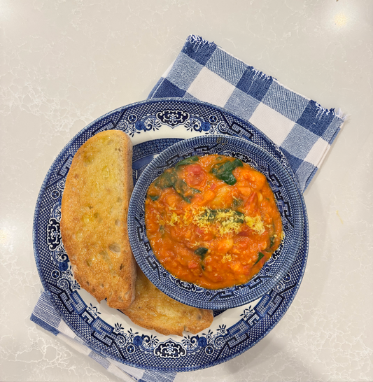 A photo of a tomato, spinach and bean stew in a blue bowl on a blue plate with toast on the side and a tea towel underneath.