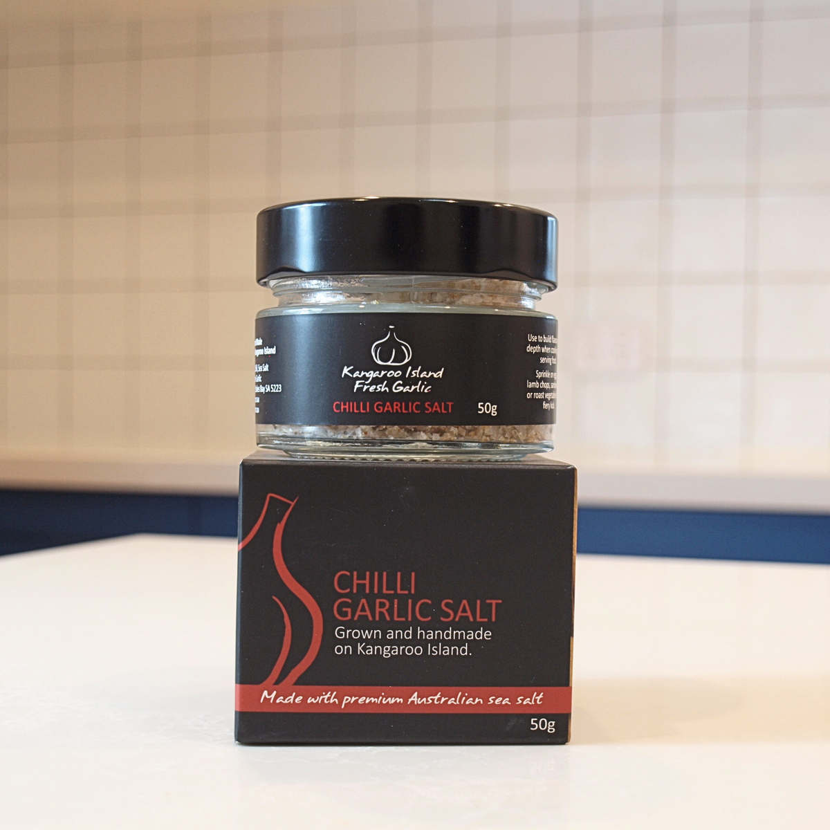 A jar of Kangaroo Island Fresh Garlic Chilli Garlic Salt with its black cardboard packaging box, set on a white countertop against a tiled kitchen background.