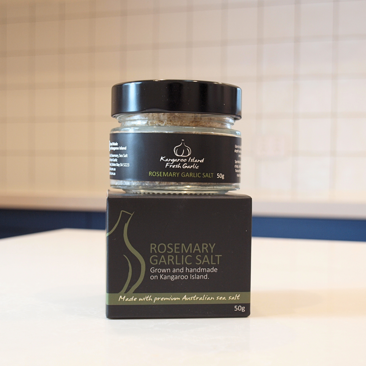 A jar of Rosemary Garlic Salt sits on its black retail box on a white countertop, with a tiled kitchen backsplash in the background.
