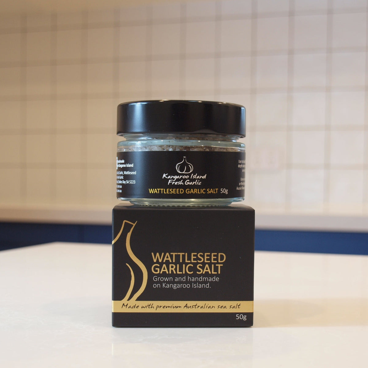 A jar of Kangaroo Island Fresh Garlic's Wattleseed Garlic Salt resting on its black and gold cardboard packaging box, set on a white countertop with a tiled kitchen background.
