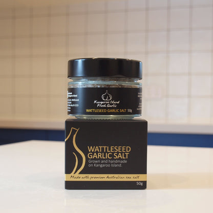 A jar of Kangaroo Island Fresh Garlic's Wattleseed Garlic Salt resting on its black and gold cardboard packaging box, set on a white countertop with a tiled kitchen background.
