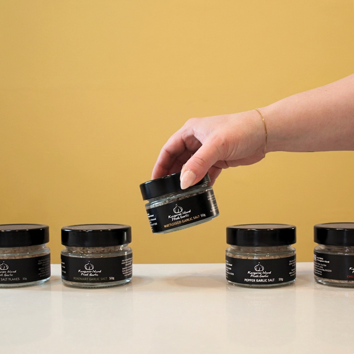 A hand holds a small jar of Wattleseed garlic Salt above a row of similar jars on a white surface against a mustard yellow background.